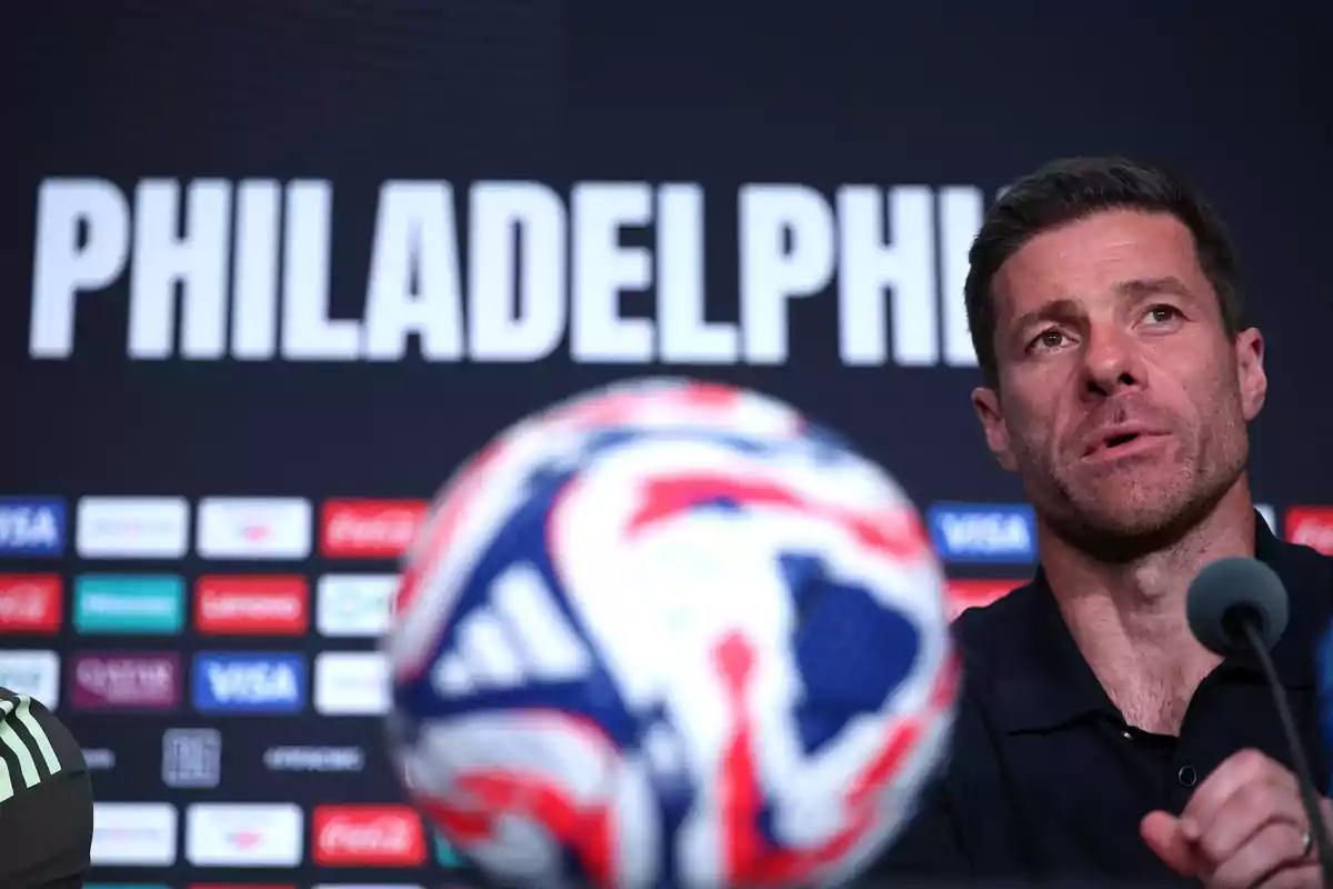 Hombre hablando en una conferencia de prensa con la palabra Philadelphia de fondo y un balón de fútbol desenfocado en primer plano
