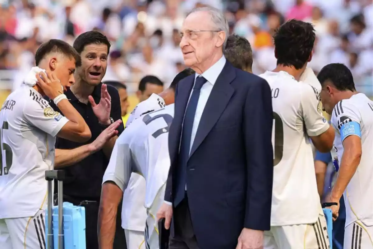 Un hombre de traje camina frente a un grupo de jugadores de fútbol vestidos de blanco que están reunidos junto a su entrenador en un estadio lleno de público.