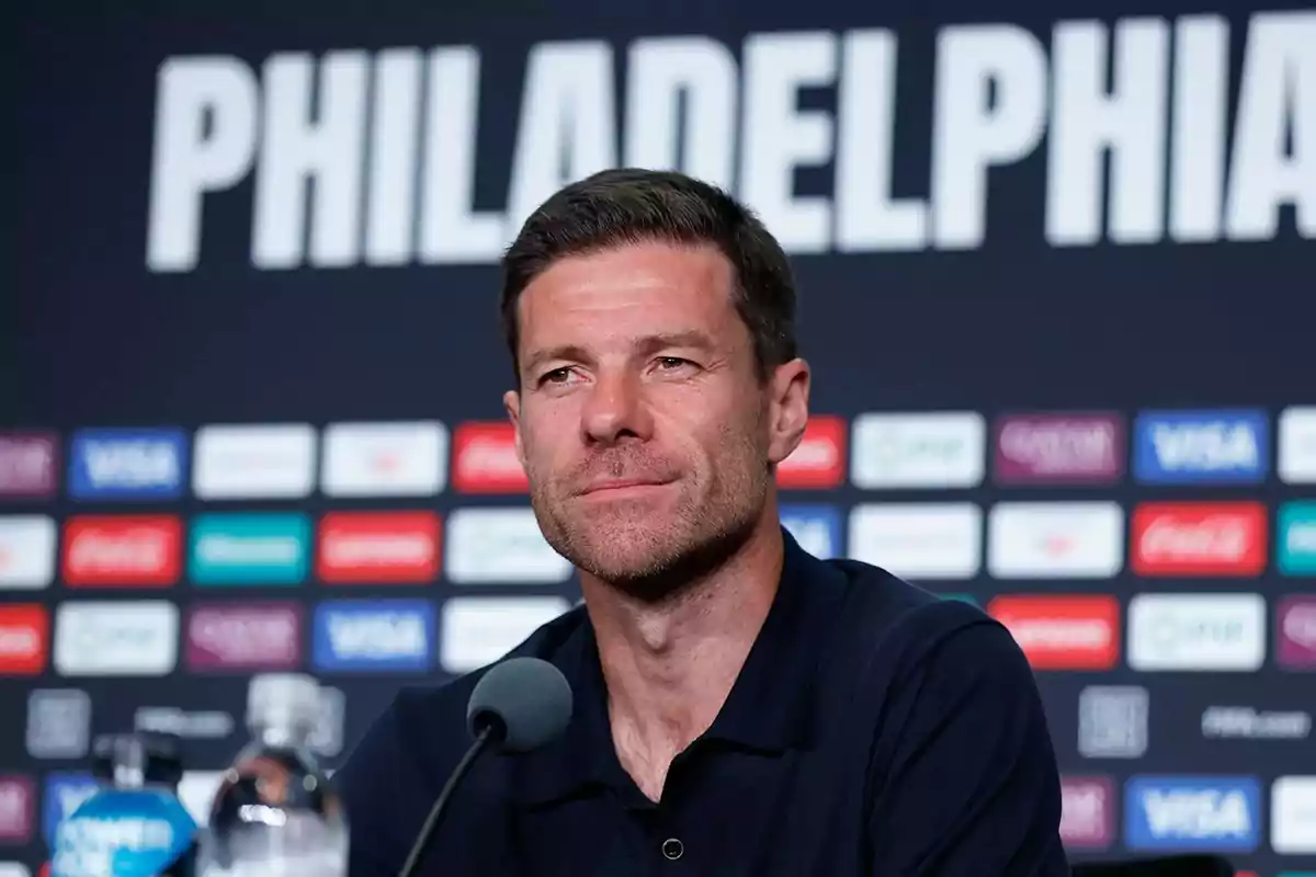 Hombre sentado frente a un micrófono en una conferencia de prensa con la palabra PHILADELPHIA de fondo y varios logotipos de patrocinadores en la pared