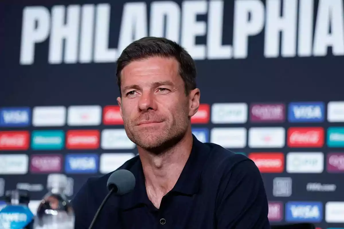 Man sitting in front of a microphone at a press conference with a background of logos and the word PHILADELPHIA behind him