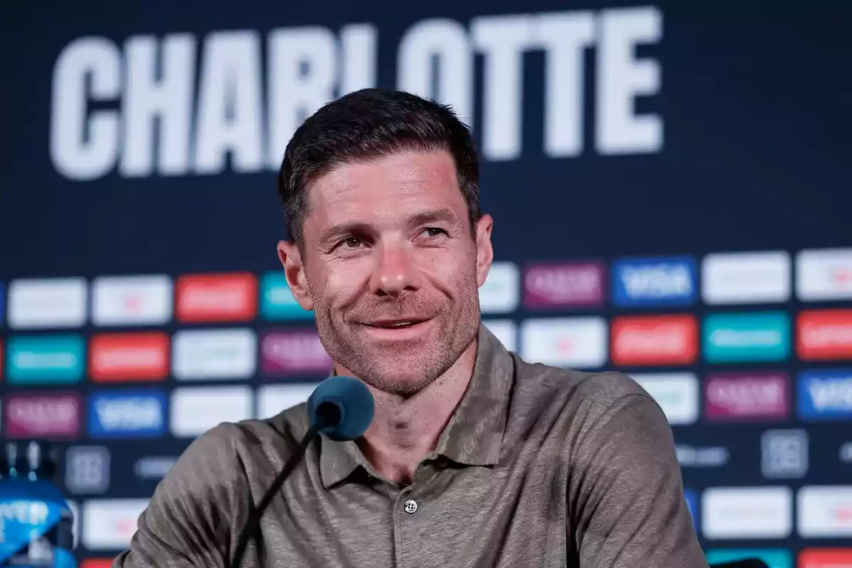 Short-haired man with a light beard speaking at a press conference with a background of logos and the word Charlotte behind him Short-haired man with a light beard speaking at a press conference with a background of logos and the word Charlotte behind him