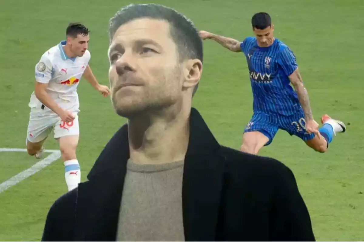 Un hombre con abrigo oscuro en primer plano y al fondo dos futbolistas en acción durante un partido en un campo de fútbol.