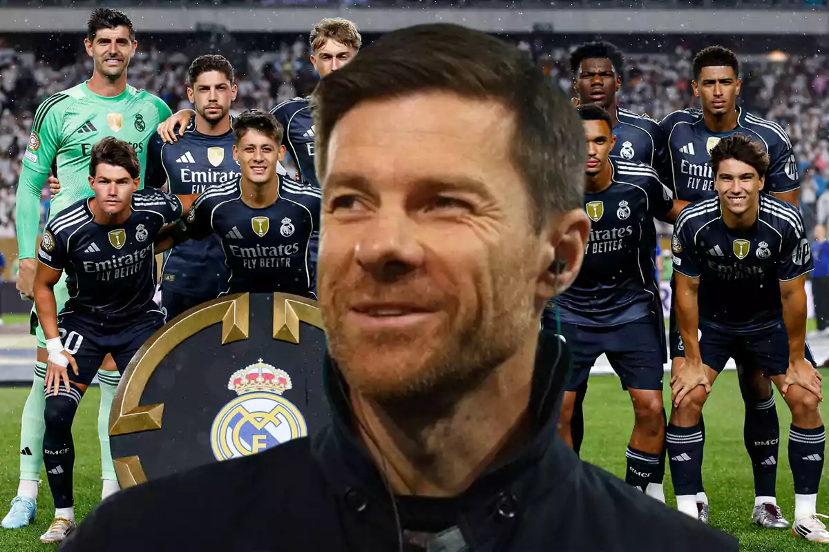 Un hombre sonriente en primer plano con un equipo de fútbol del Real Madrid posando en el fondo sobre el campo de juego