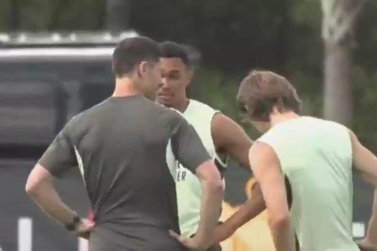 Three people are talking on a sports training field; two of them are wearing light-colored sleeveless jerseys and the other is wearing a dark jersey.
