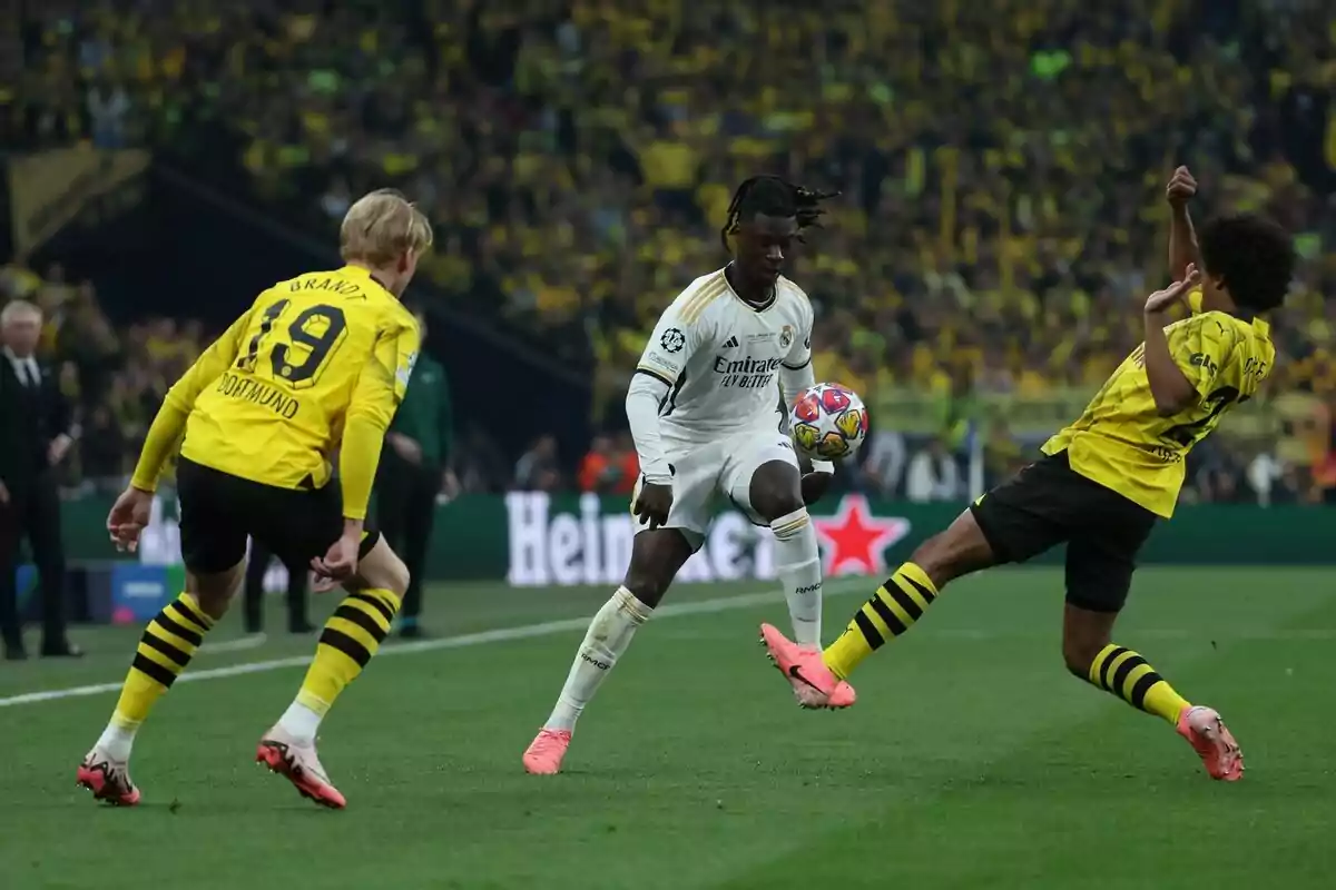 Un jugador del Real Madrid controla el balón mientras dos futbolistas del Borussia Dortmund intentan quitarle la pelota durante un partido de fútbol en un estadio lleno de aficionados. Un jugador del Real Madrid controla el balón mientras dos futbolistas del Borussia Dortmund intentan quitarle la pelota durante un partido de fútbol en un estadio lleno de aficionados.