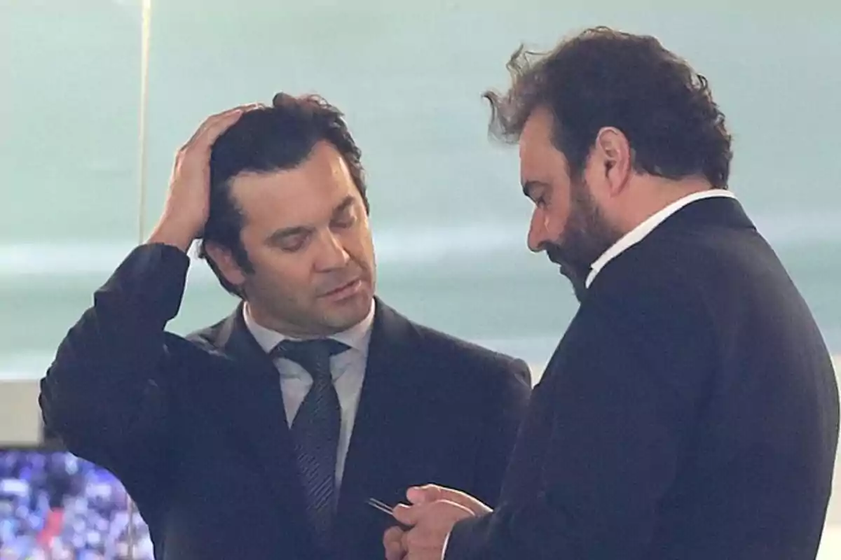 Dos hombres de traje conversan, uno de ellos se toca la cabeza mientras el otro sostiene un objeto pequeño en las manos Dos hombres de traje conversan, uno de ellos se toca la cabeza mientras el otro sostiene un objeto pequeño en las manos