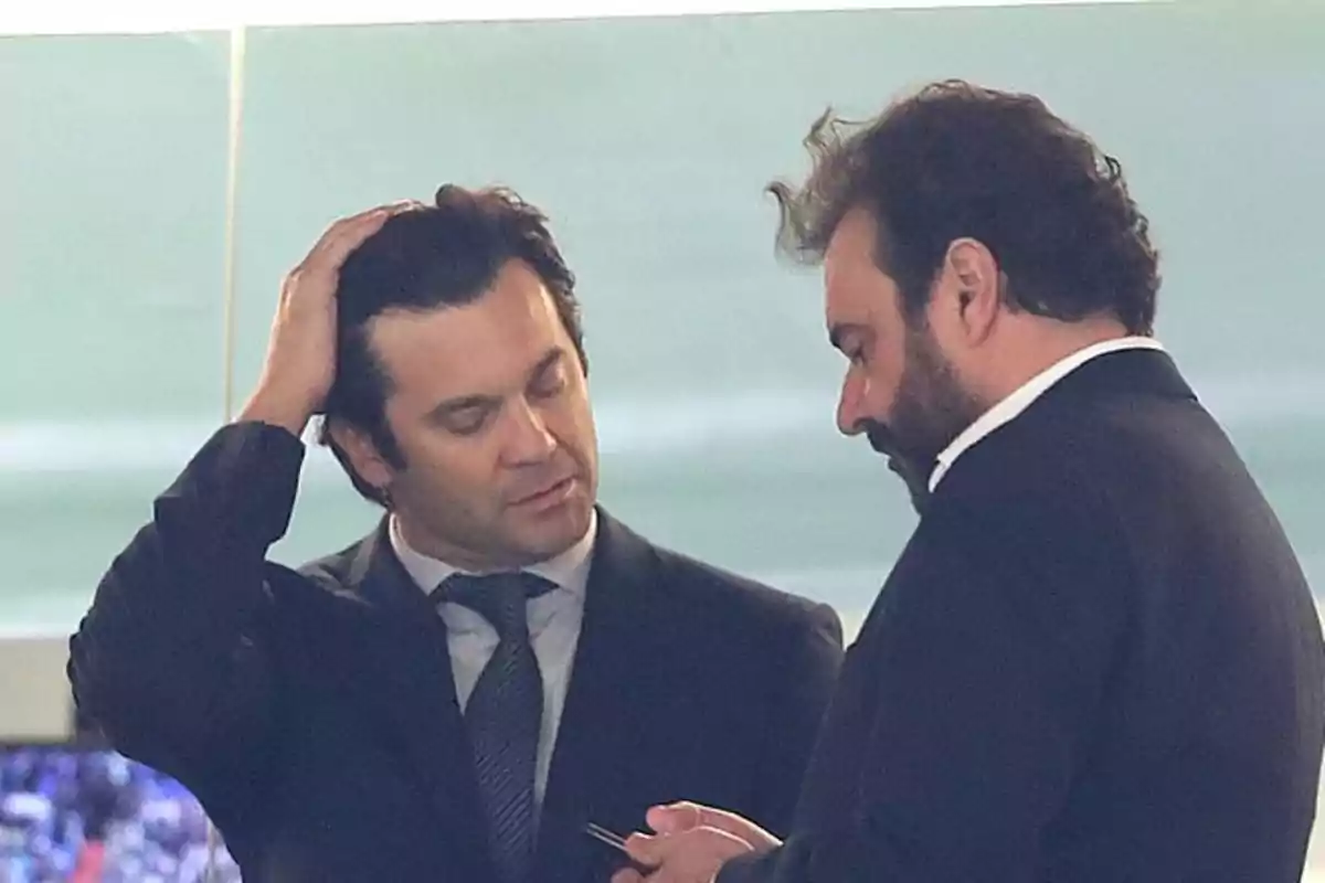 Dos hombres de traje conversan en un ambiente formal mientras uno de ellos se toca la cabeza y el otro sostiene un objeto en las manos Dos hombres de traje conversan en un ambiente formal mientras uno de ellos se toca la cabeza y el otro sostiene un objeto en las manos