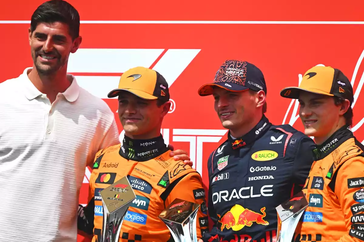 Four people pose smiling in front of a red background; three of them are wearing racing uniforms and caps, holding silver trophies. Four people pose smiling in front of a red background; three of them are wearing racing uniforms and caps, holding silver trophies.