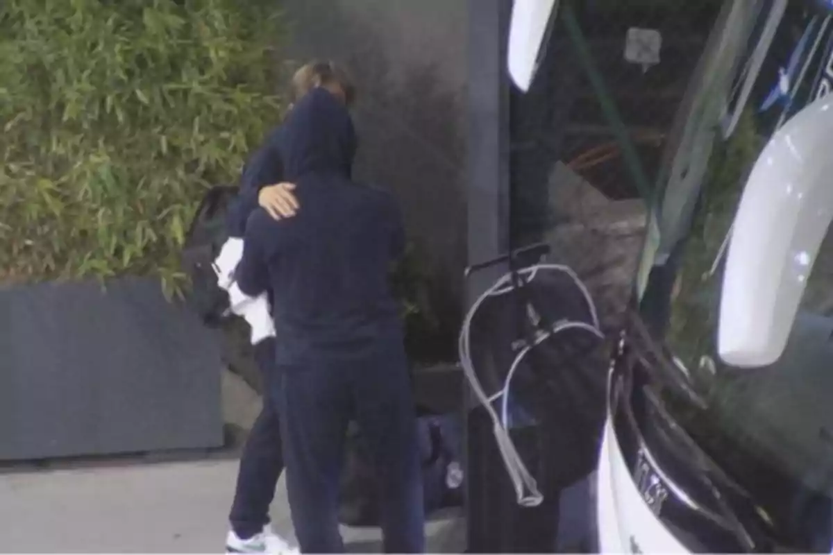 Two people are hugging next to a parked bus and a plant in the background. Two people are hugging next to a parked bus and a plant in the background.