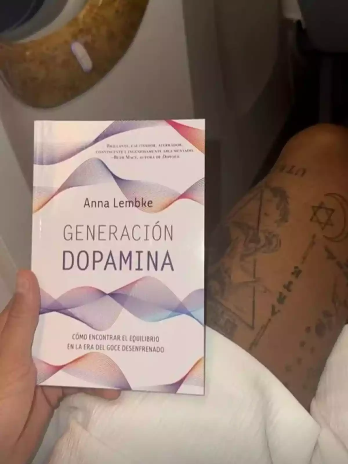 Person holding the book Dopamine Nation by Anna Lembke while sitting, showing tattoos on their leg Person holding the book Dopamine Nation by Anna Lembke while sitting, showing tattoos on their leg