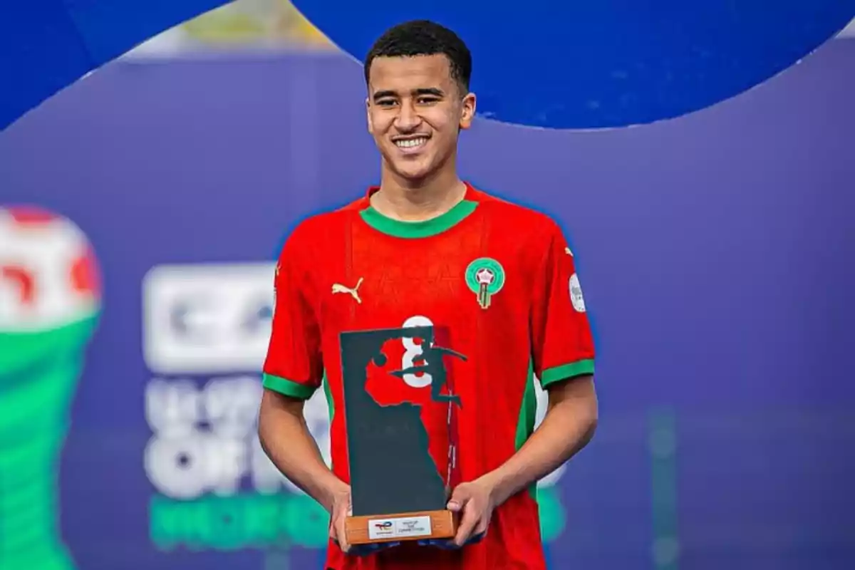 player in a red Morocco uniform smiles while holding a black rectangular trophy with the silhouette of the African continent and a player figure. player in a red Morocco uniform smiles while holding a black rectangular trophy with the silhouette of the African continent and a player figure.