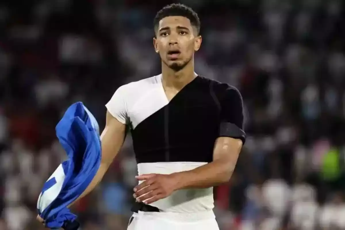 Futbolista en el campo sosteniendo una camiseta azul con expresión de cansancio y usando una prenda deportiva ajustada en blanco y negro Futbolista en el campo sosteniendo una camiseta azul con expresión de cansancio y usando una prenda deportiva ajustada en blanco y negro