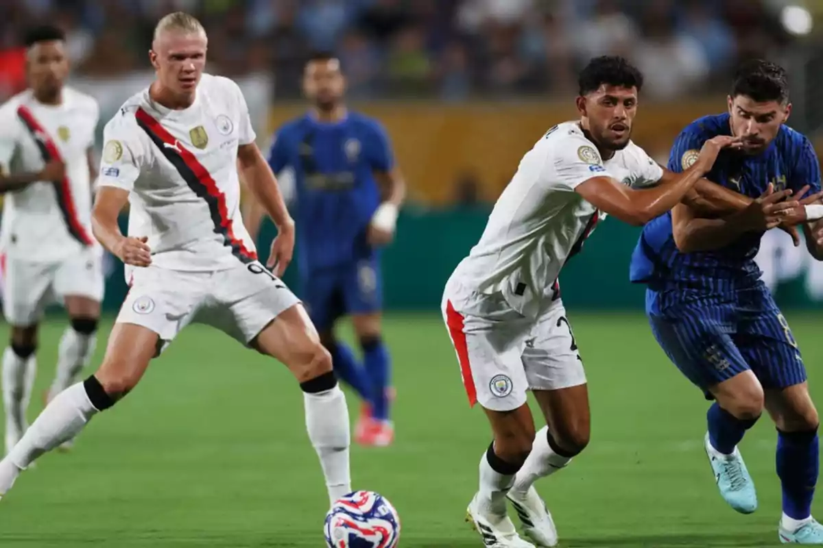Jugadores de fútbol disputan el balón durante un partido entre un equipo con uniforme blanco y otro con uniforme azul en un estadio Jugadores de fútbol disputan el balón durante un partido entre un equipo con uniforme blanco y otro con uniforme azul en un estadio