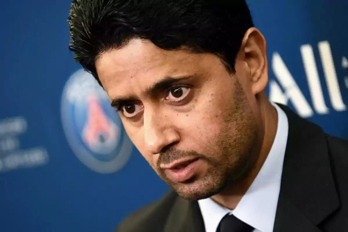 Hombre de cabello oscuro y barba corta con expresión seria usando traje frente a un fondo azul con el logo de un equipo deportivo desenfocado.