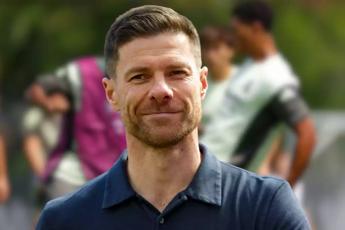 Hombre de cabello corto y barba ligera sonríe mientras está al aire libre con personas desenfocadas en el fondo
