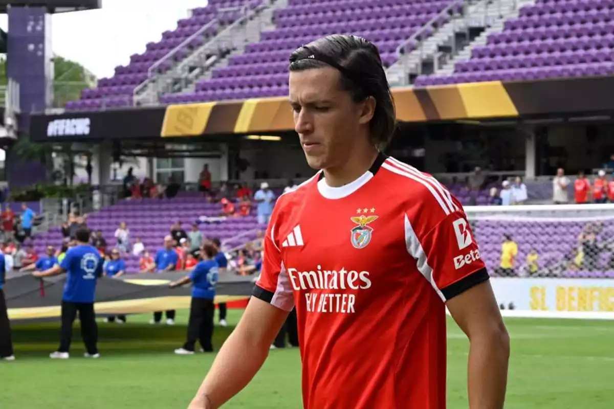 Futbolista con camiseta roja del Benfica caminando en un estadio con gradas moradas y personas al fondo Futbolista con camiseta roja del Benfica caminando en un estadio con gradas moradas y personas al fondo