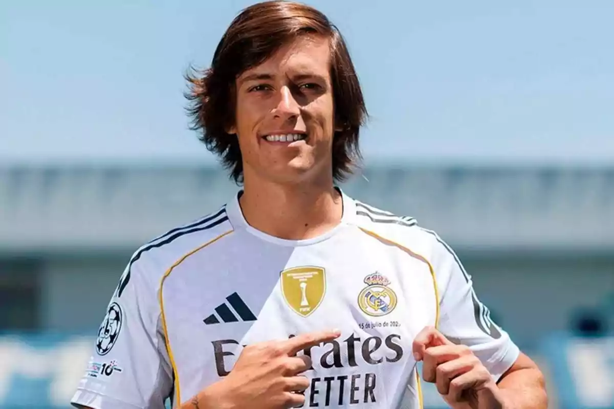Joven con cabello largo y castaño sonriendo y señalando el escudo de un equipo de fútbol en su camiseta blanca
