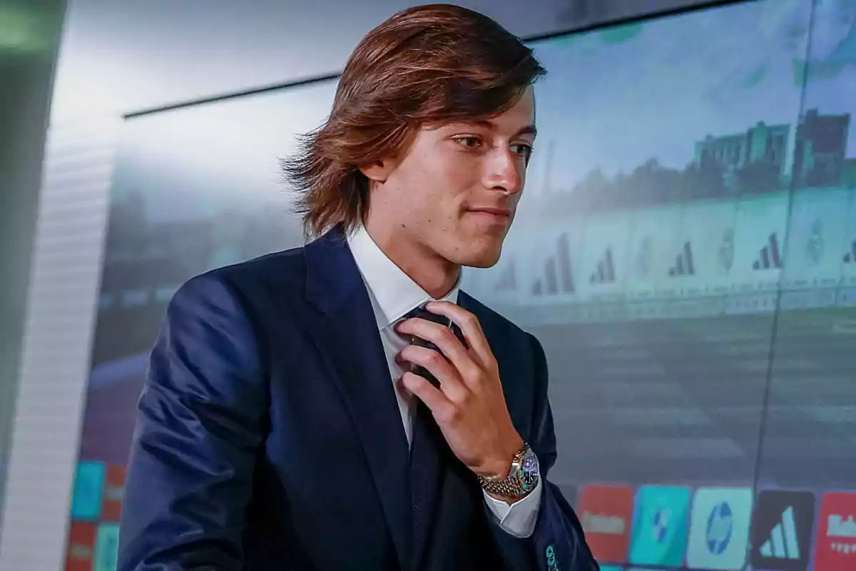 Joven con traje azul ajustándose la corbata frente a una pantalla con gráficos y logotipos.
