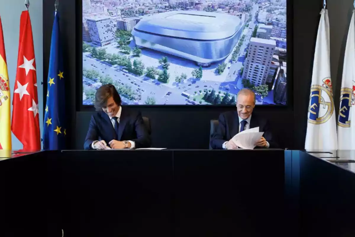 Dos hombres con traje firmando documentos en una mesa frente a banderas y una pantalla que muestra una imagen aérea de un estadio moderno