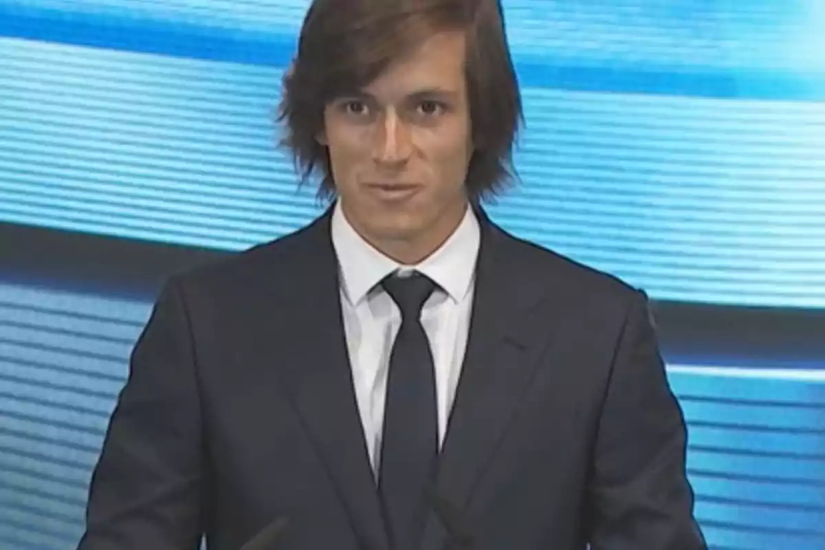 Young man with long brown hair wearing a dark suit and tie in front of a blue background
