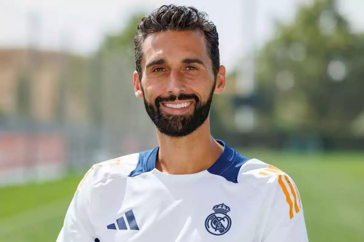 Hombre con barba y cabello corto rizado sonríe mientras viste una camiseta blanca del Real Madrid en un campo de entrenamiento al aire libre