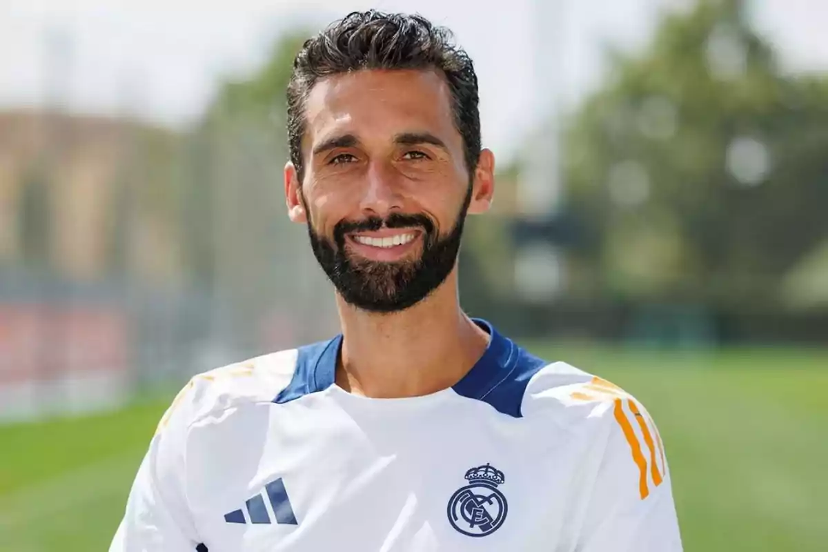 Hombre con barba y cabello oscuro sonríe mientras viste una camiseta blanca del Real Madrid en un campo de entrenamiento al aire libre Hombre con barba y cabello oscuro sonríe mientras viste una camiseta blanca del Real Madrid en un campo de entrenamiento al aire libre
