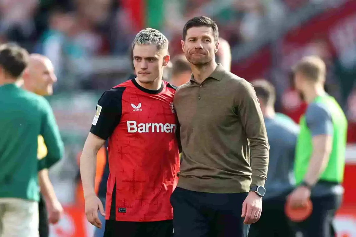 Dos personas en un campo de fútbol, una con uniforme rojo y negro y otra con ropa casual, rodeadas de otras personas desenfocadas al fondo. Dos personas en un campo de fútbol, una con uniforme rojo y negro y otra con ropa casual, rodeadas de otras personas desenfocadas al fondo.