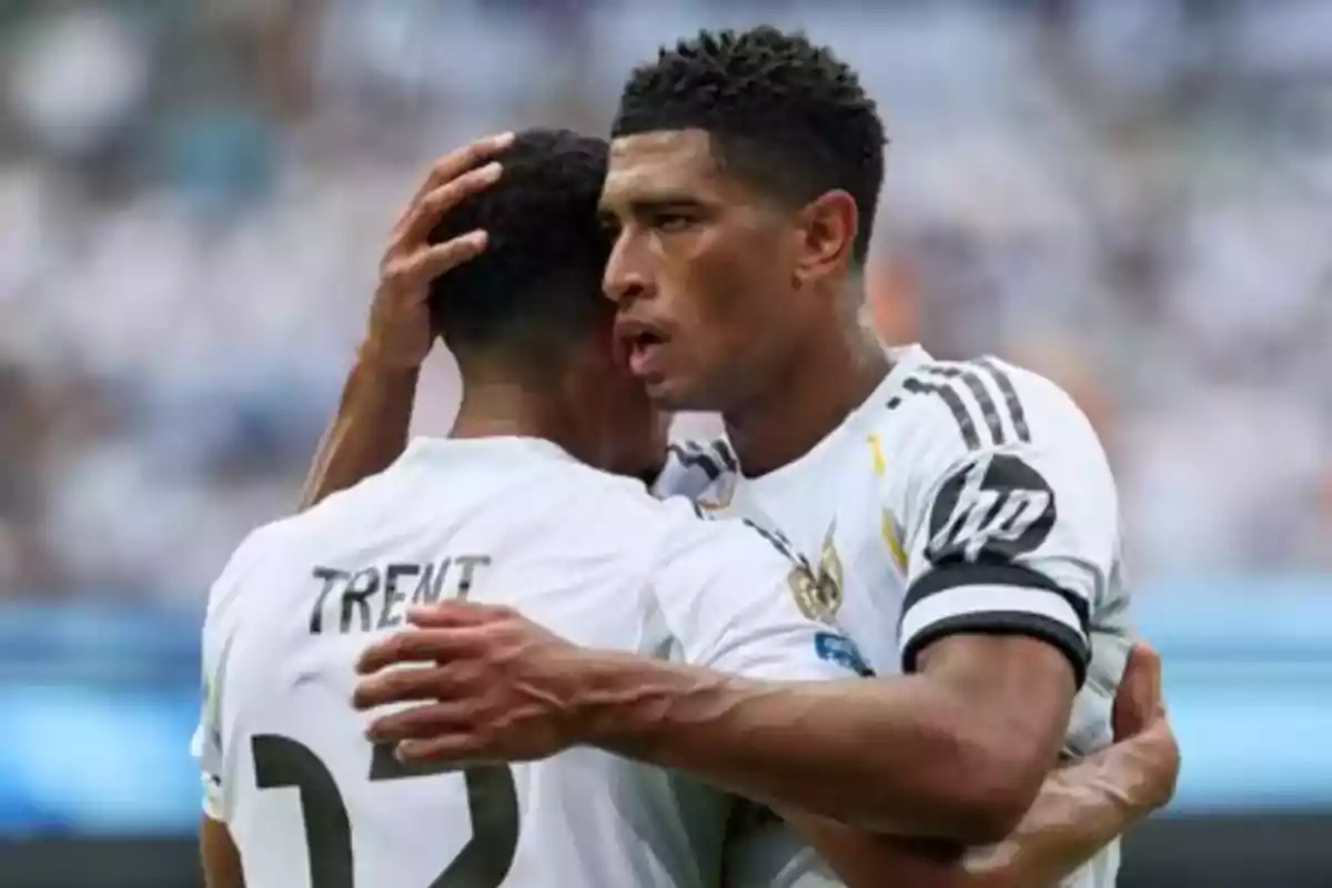 Dos futbolistas con uniforme blanco se abrazan en el campo durante un partido