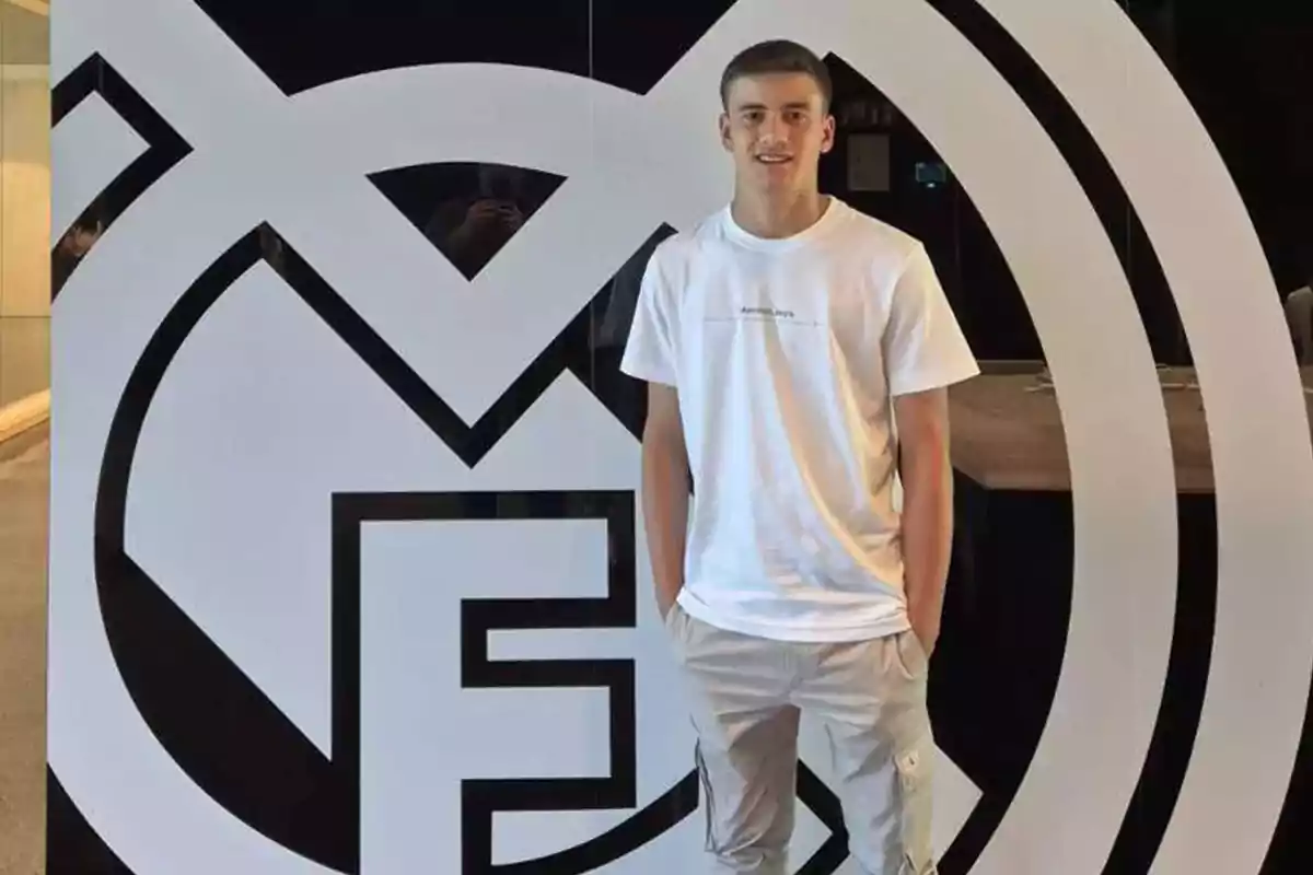 Joven con camiseta blanca y pantalón claro posando frente a un gran escudo del Real Madrid Joven con camiseta blanca y pantalón claro posando frente a un gran escudo del Real Madrid