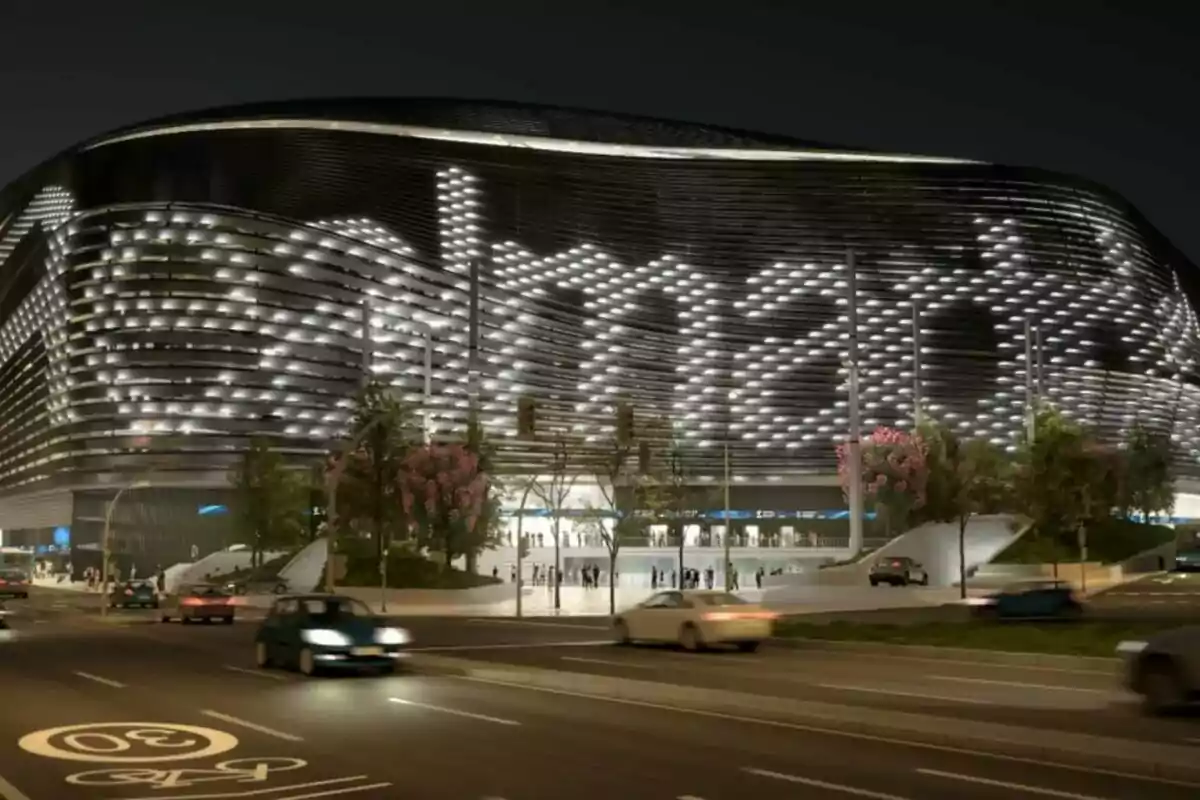 Fachada iluminada de un estadio moderno con autos circulando por la calle frente a él durante la noche Fachada iluminada de un estadio moderno con autos circulando por la calle frente a él durante la noche