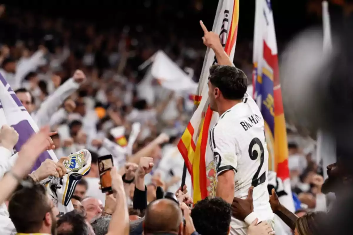Jugador de fútbol celebrando con la afición en las gradas mientras sostiene una bandera y la multitud lo rodea con entusiasmo Jugador de fútbol celebrando con la afición en las gradas mientras sostiene una bandera y la multitud lo rodea con entusiasmo