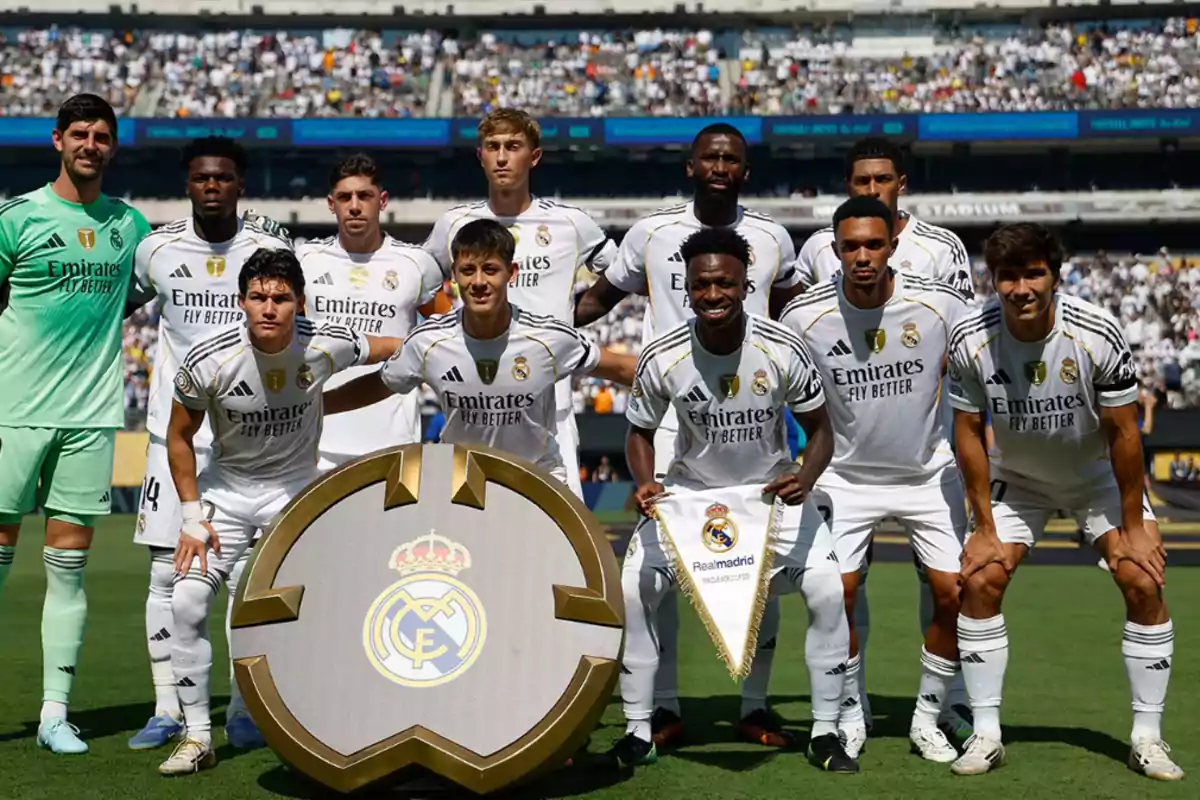 Once jugadores del Real Madrid posan en el campo antes de un partido con el escudo del club al frente y el estadio lleno de aficionados al fondo Once jugadores del Real Madrid posan en el campo antes de un partido con el escudo del club al frente y el estadio lleno de aficionados al fondo