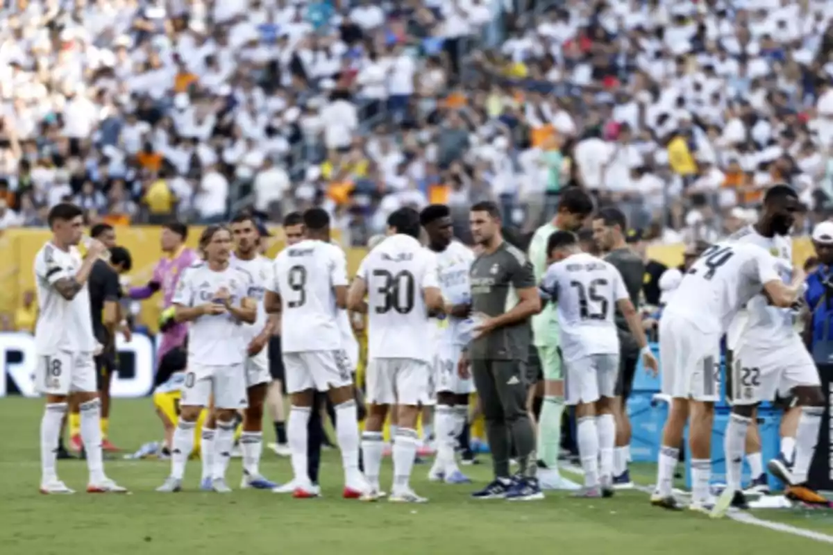 Jugadores de un equipo de fútbol vestidos de blanco se agrupan en el campo durante un partido con el público en las gradas al fondo Jugadores de un equipo de fútbol vestidos de blanco se agrupan en el campo durante un partido con el público en las gradas al fondo