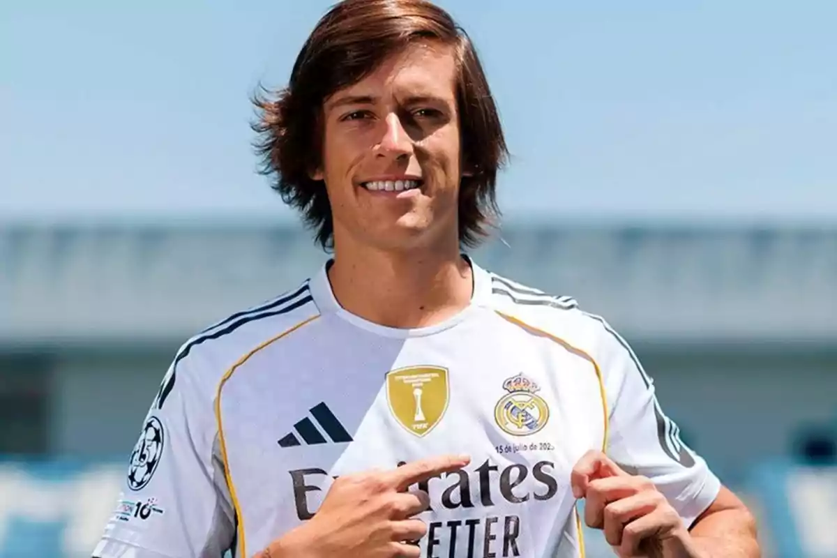 Joven con cabello castaño y largo sonríe mientras señala el escudo del Real Madrid en su camiseta blanca
