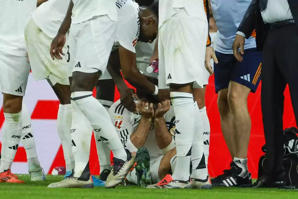 Jugadores de fútbol rodean y consuelan a un compañero que está sentado en el césped cubriéndose el rostro con las manos durante un partido