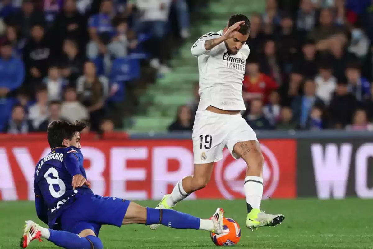 Un jugador de fútbol con uniforme blanco esquiva una barrida de un rival vestido de azul durante un partido en un estadio lleno de espectadores.