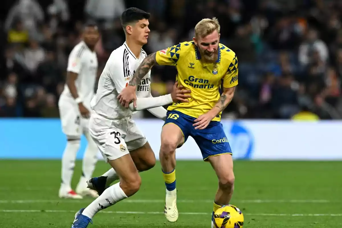 Two players compete for the ball during a match, one wearing a white uniform and the other wearing a yellow and blue uniform. Two players compete for the ball during a match, one wearing a white uniform and the other wearing a yellow and blue uniform.