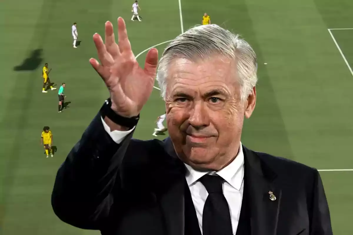 Entrenador de fútbol con traje oscuro saludando con la mano en alto sobre el fondo de un campo de fútbol donde se juega un partido