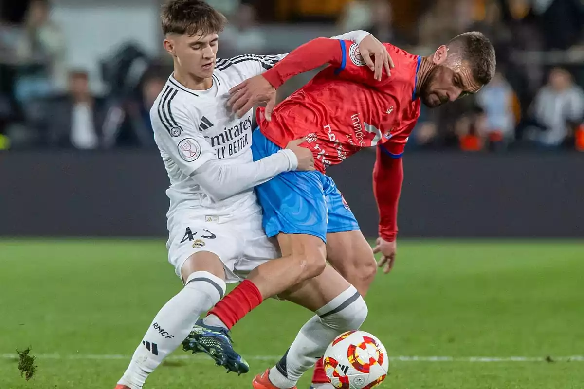 Dos futbolistas disputan el balón durante un partido uno con uniforme blanco y el otro con uniforme rojo y azul Dos futbolistas disputan el balón durante un partido uno con uniforme blanco y el otro con uniforme rojo y azul