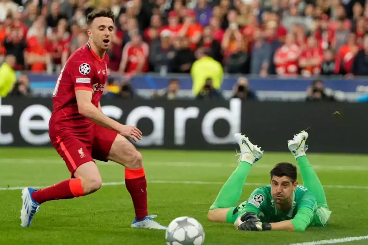 Un jugador de fútbol con uniforme rojo observa mientras el portero rival, vestido de verde, está en el suelo tras intentar detener el balón durante un partido importante con público en las gradas. Un jugador de fútbol con uniforme rojo observa mientras el portero rival, vestido de verde, está en el suelo tras intentar detener el balón durante un partido importante con público en las gradas.