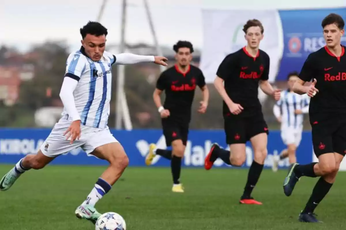 Jugador con uniforme blanco y azul controla el balón mientras es perseguido por varios rivales con uniforme negro en un partido de fútbol en campo abierto