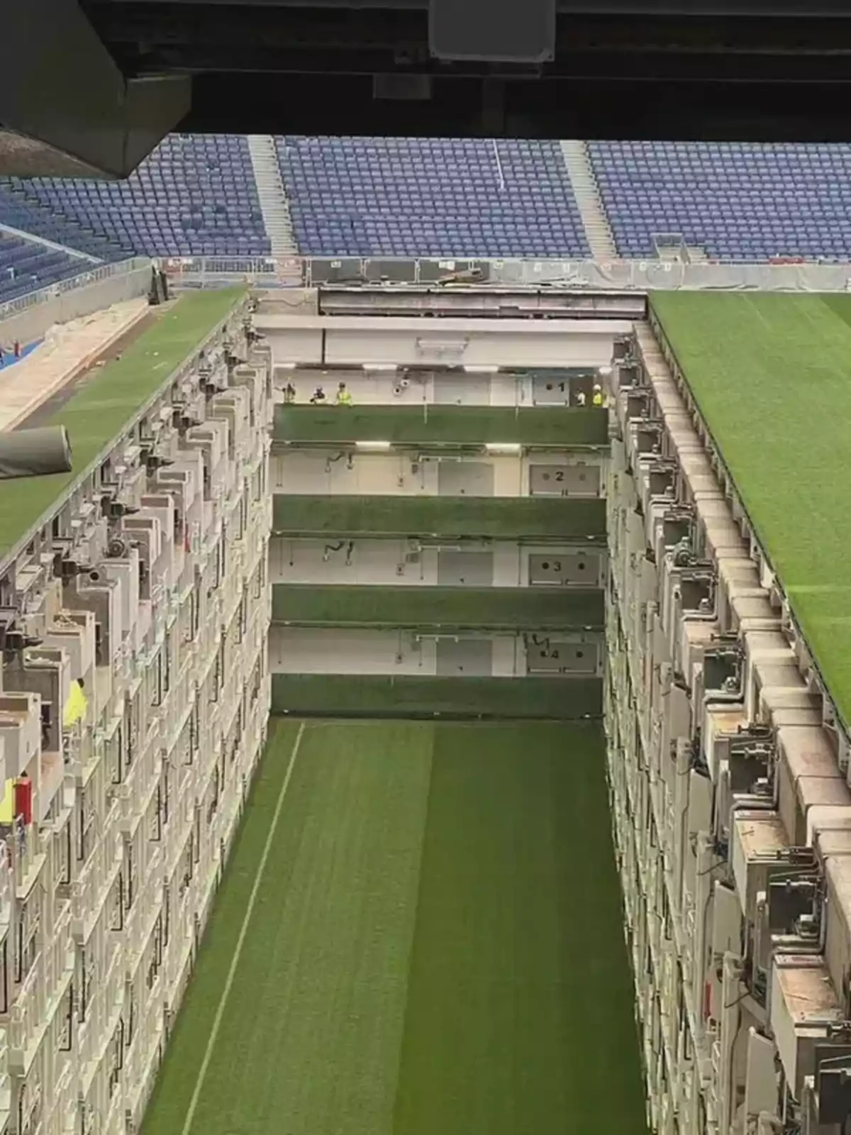 Vista de un estadio con gradas azules y una sección del campo de fútbol que se desliza hacia adentro, mostrando el mecanismo subterráneo que permite mover el césped Vista de un estadio con gradas azules y una sección del campo de fútbol que se desliza hacia adentro, mostrando el mecanismo subterráneo que permite mover el césped