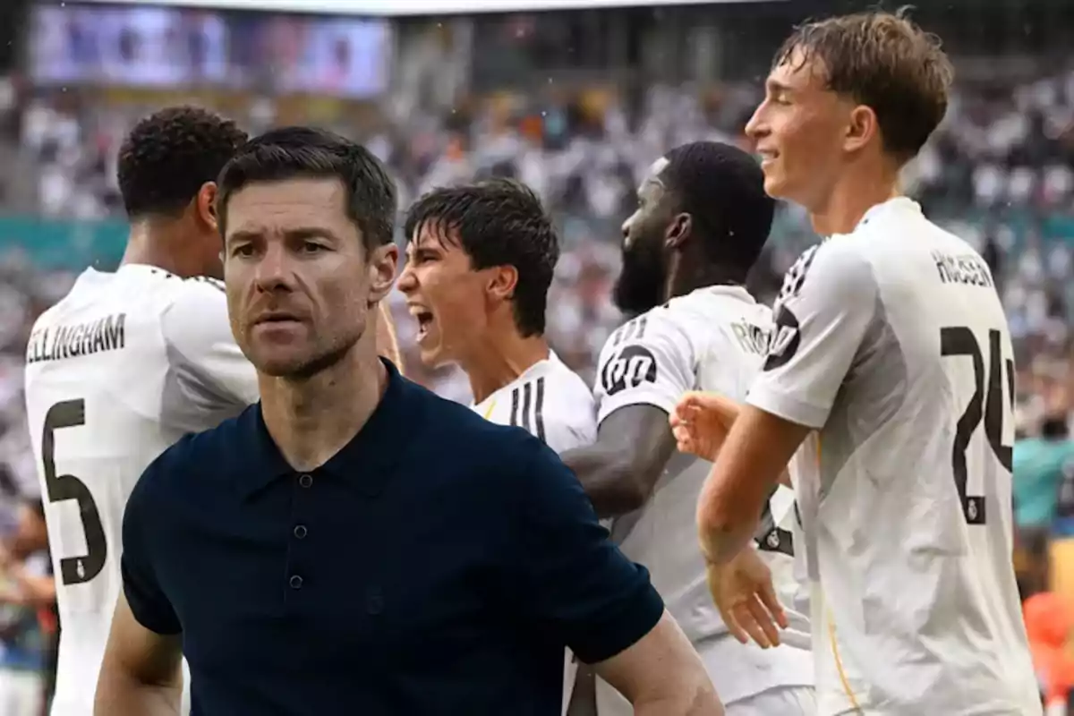 Un entrenador de fútbol con expresión seria al frente mientras varios jugadores de un equipo celebran en el fondo durante un partido