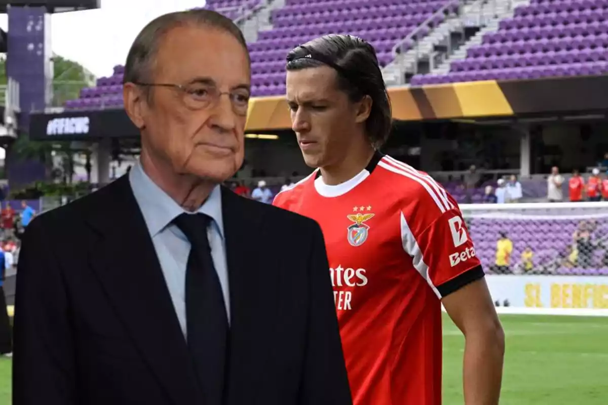 A man in a dark suit is in the foreground while in the background a player wearing Benfica’s red uniform walks in a stadium with purple stands.