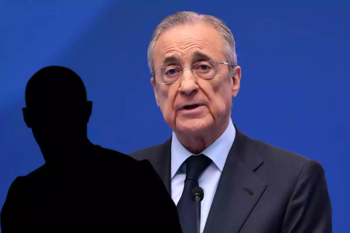 Older man in a suit and tie speaking into a microphone with a black silhouette of a person in the foreground against a blue background