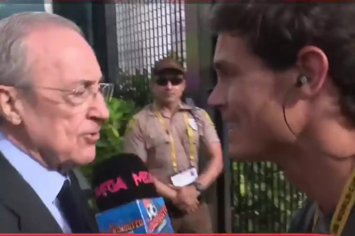 Un hombre mayor con gafas y traje es entrevistado por un reportero que sostiene un micrófono negro con letras rosas mientras un guardia de seguridad observa al fondo