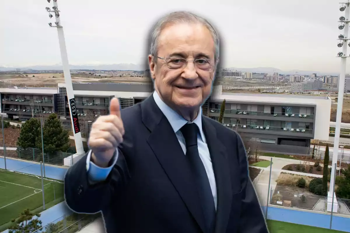 Older man with glasses and a suit smiles and gives a thumbs up in front of a modern sports complex