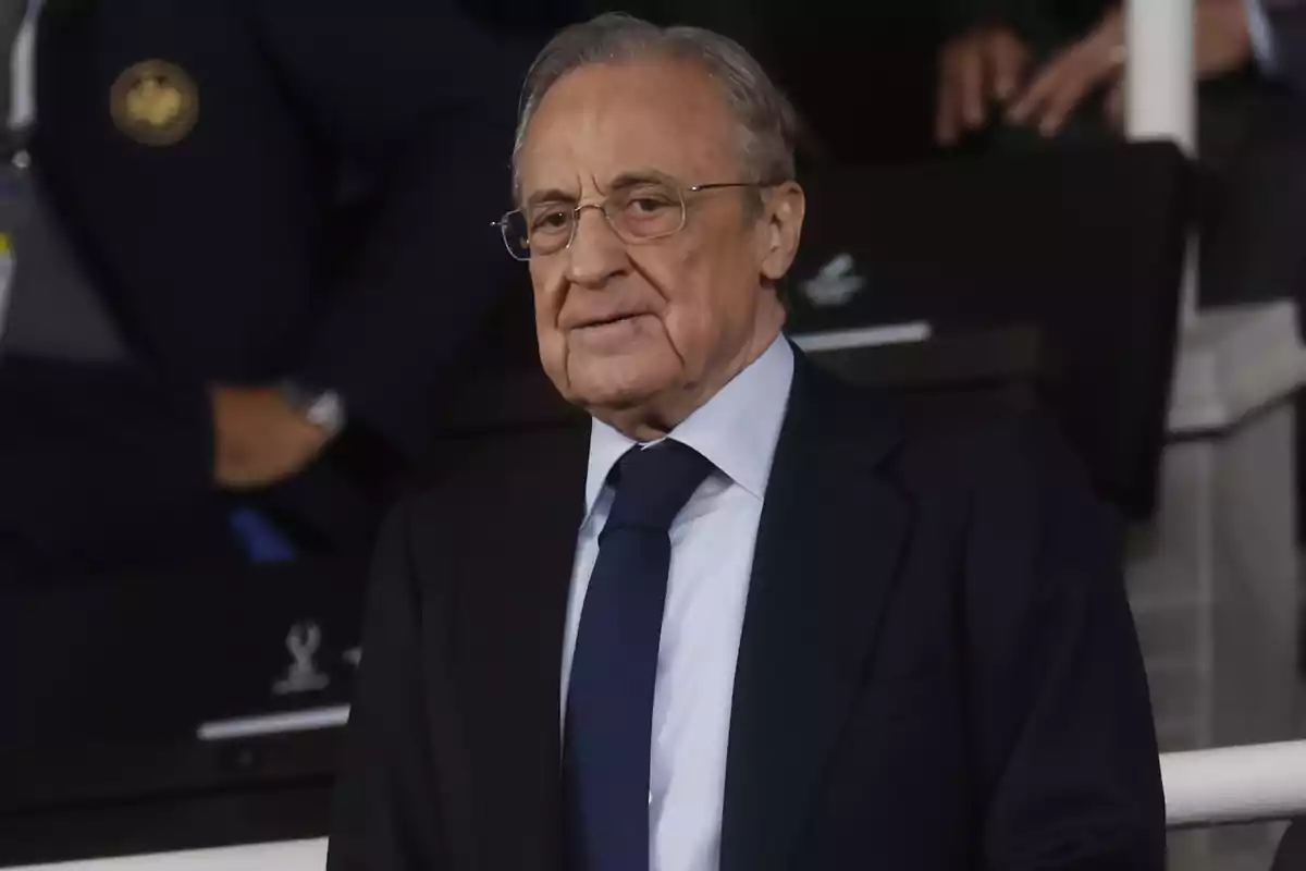 Older man with gray hair and glasses wearing a dark suit and blue tie at a formal event