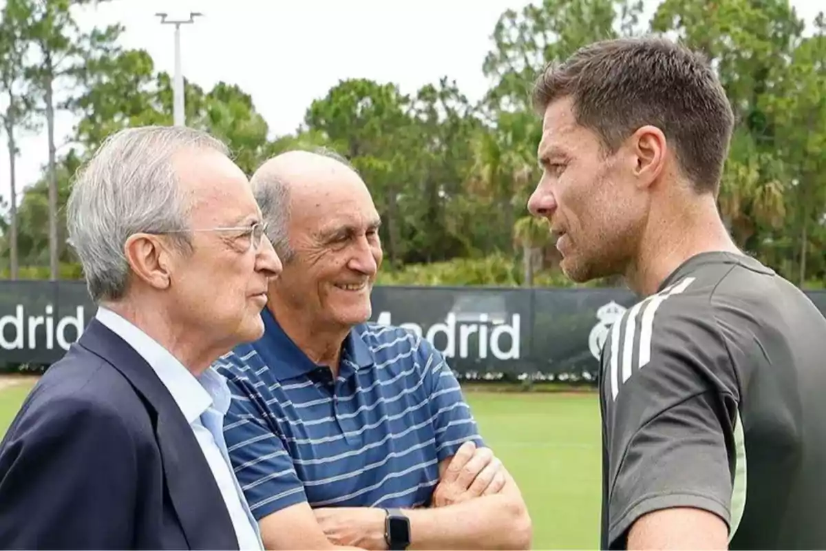 Tres hombres conversan en un campo de entrenamiento de fútbol con árboles al fondo y un cartel de Madrid parcialmente visible