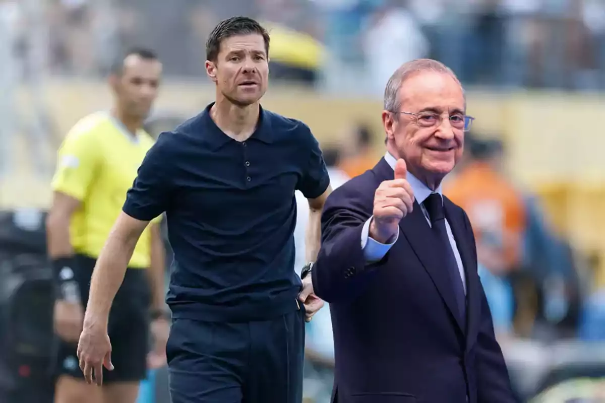 A man in a dark blue jersey on a soccer field and another older man in a suit and tie giving a thumbs-up gesture.